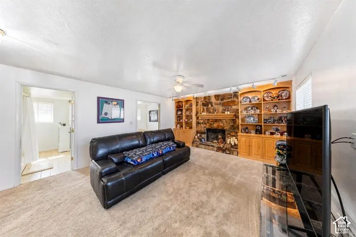 Carpeted living area featuring a stone fireplace with wood mantel, built-in shelving, wood cabinetry, and a ceiling fan
