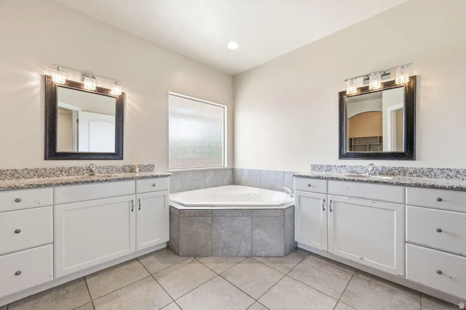 Spa-inspired primary bathroom showcasing a luxurious corner soaking tub with tile surround, dual vanities with white cabinetry and granite countertops, framed mirrors with integrated lighting, and neutral tile flooring throughout