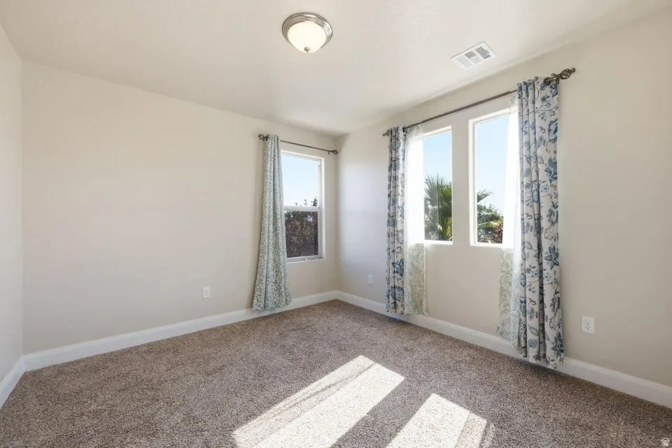 Bright corner bedroom featuring neutral carpet flooring, dual windows with blue patterned curtains, and abundant natural light