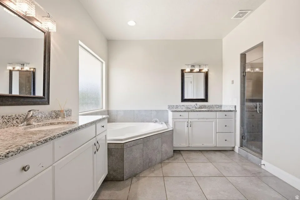 Spacious bathroom featuring a corner soaking tub with tile surround, a glass-enclosed shower, dual vanities with light-toned granite countertops, and tile flooring