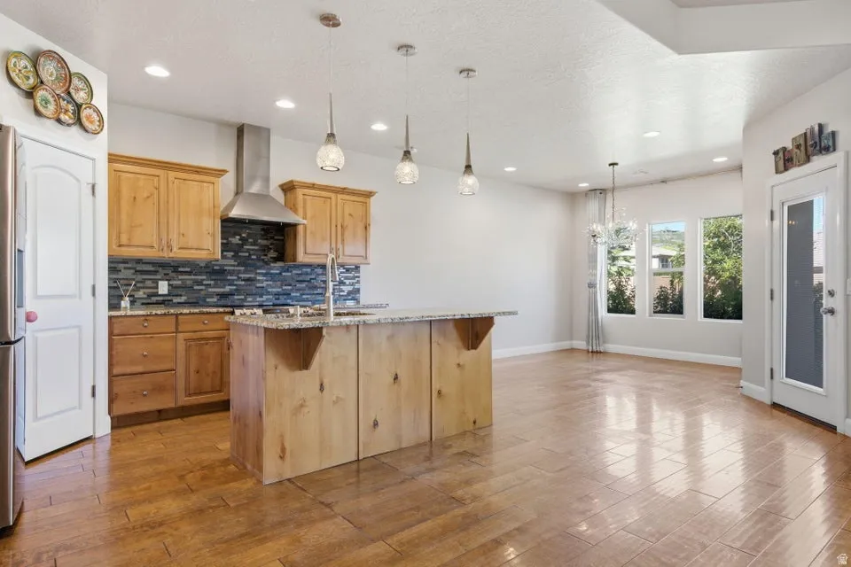 Open-concept kitchen and dining area featuring warm wood cabinetry, a large center island with wood-finish base, stainless steel pendant lighting, and a blue tile backsplash
