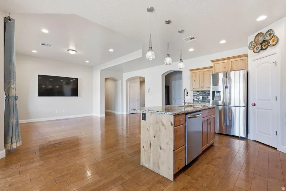 Open-concept kitchen and living area featuring wood-finish flooring throughout, a large center island with white cabinetry and wood-tone accents, stainless steel appliances including a refrigerator and dishwasher, pendant lighting, recessed ceiling fixtur