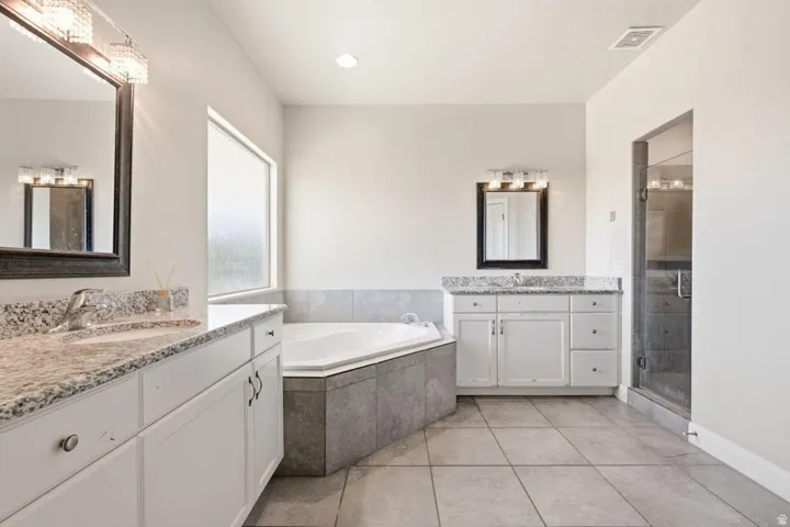Spacious bathroom featuring a corner soaking tub with tile surround, a glass-enclosed shower, dual vanities with light-toned granite countertops, and tile flooring