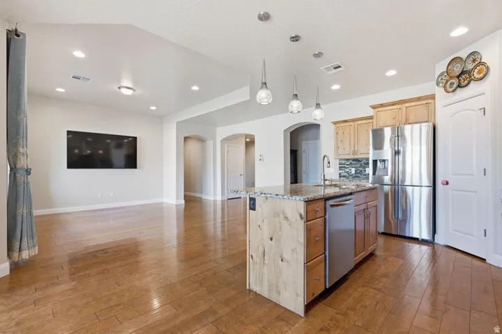 Open-concept kitchen and living area featuring wood-finish flooring throughout, a large center island with white cabinetry and wood-tone accents, stainless steel appliances including a refrigerator and dishwasher, pendant lighting, recessed ceiling fixtur
