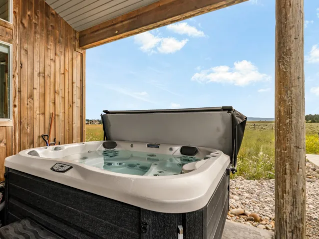 Covered deck with hot tub featuring jets and integrated seating, wood-finish siding on the structure, large support posts, metal roof overhang, and expansive open landscape with clear sightlines across the property grounds
