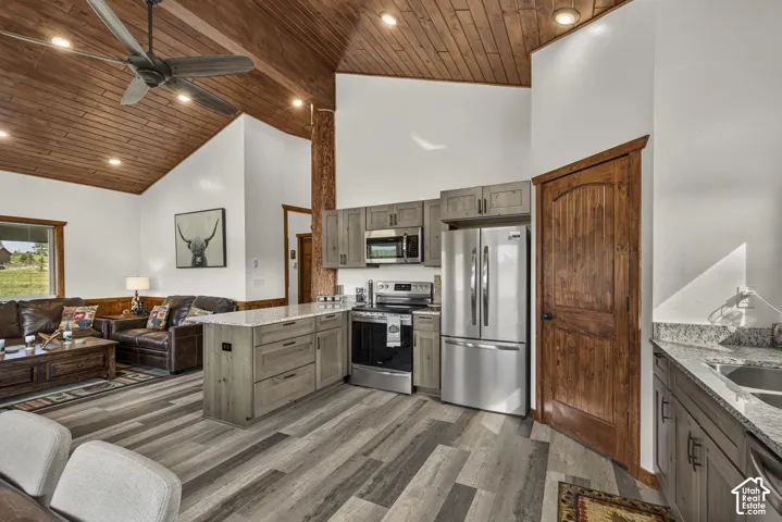 Open-concept kitchen and living area showcasing vaulted wood-finish ceilings with exposed beams, stainless steel appliances including a French-door refrigerator and built-in microwave, gray wood-tone flooring throughout, and wood cabinetry with ample stor