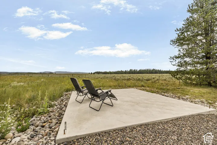 Expansive outdoor entertaining patio featuring a concrete pad with river rock bordering, positioned to overlook sprawling grassland and mature conifer trees