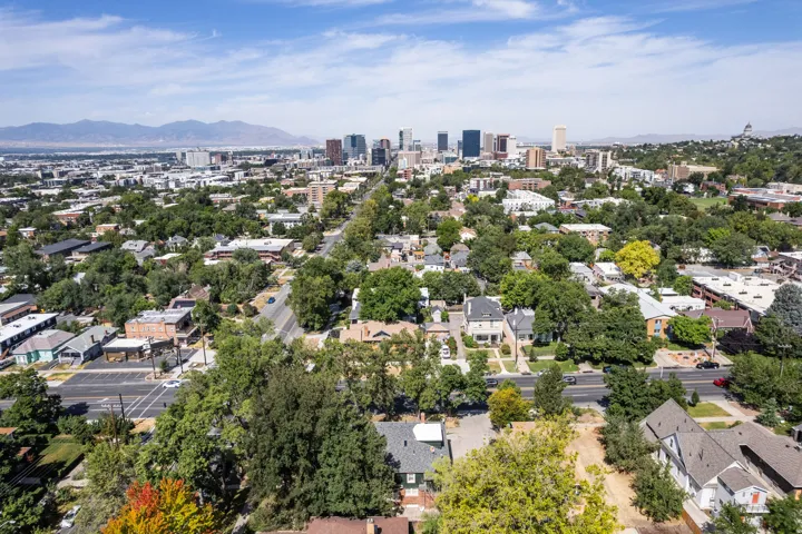Urban landscape featuring a central business district skyline, dense residential areas, mature trees, and distant mountains