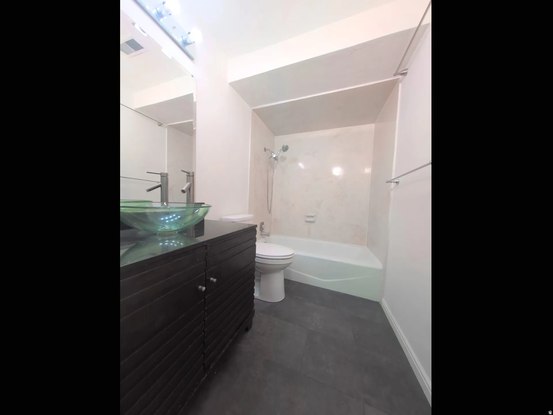 Bathroom featuring a vessel sink with a contemporary faucet, dark wood-finish vanity, dark tile flooring, and a shower-tub combination with light-toned wall surround