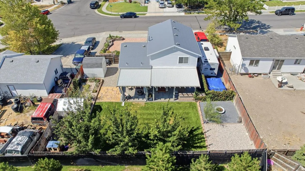 Aerial view showcasing a contemporary residence with a charcoal-gray pitched roof and white exterior walls, set on a generous lot featuring mature landscaping, a manicured lawn, brick hardscaping, and established trees providing natural privacy screening