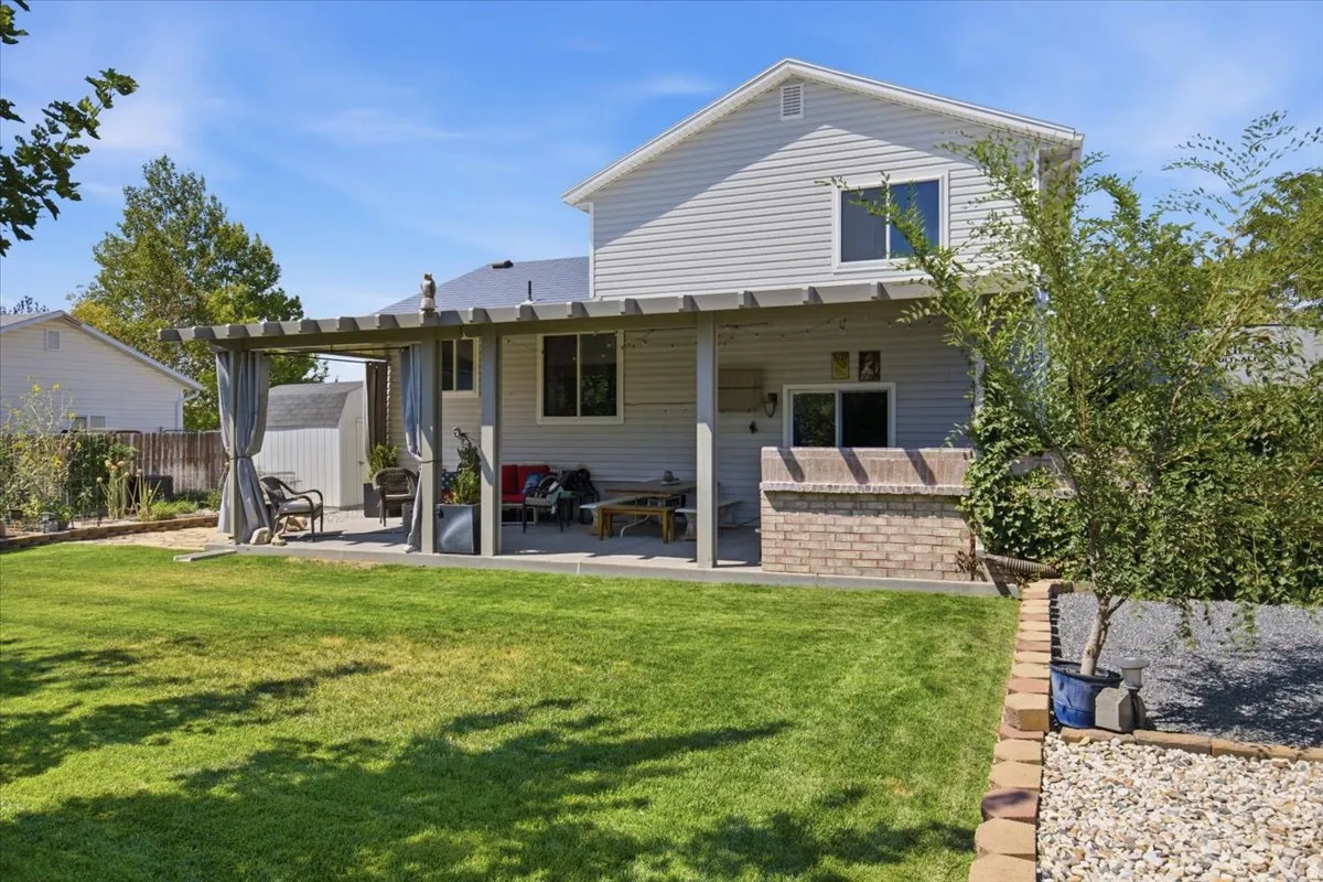 Spacious backyard oasis featuring a manicured lawn with mature landscaping, brick paver pathway, and a covered patio extension with pergola-style overhead structure