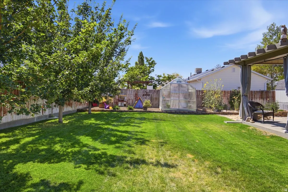 Expansive backyard featuring a large lawn, mature trees, privacy fencing, a greenhouse, and a covered patio with draped sides