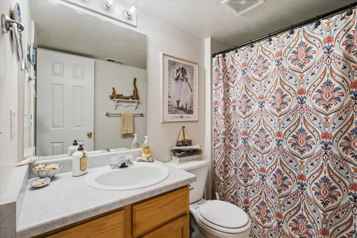 Bright bathroom featuring a white porcelain sink set in a wood-finish vanity with ample counter space, large framed mirror with integrated lighting, white subway tile backsplash, and decorative patterned shower curtain