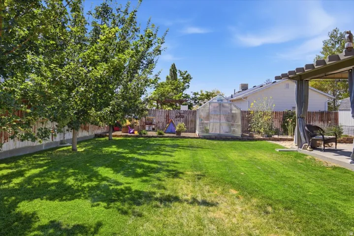 Expansive backyard featuring a large lawn, mature trees, privacy fencing, a greenhouse, and a covered patio with draped sides