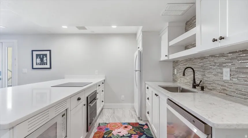 Galley kitchen featuring white shaker cabinetry, stainless steel appliances including a refrigerator and built-in range, quartz countertops, a tile backsplash, undermount sink with matte black faucet, recessed lighting, and light wood-finish flooring thro