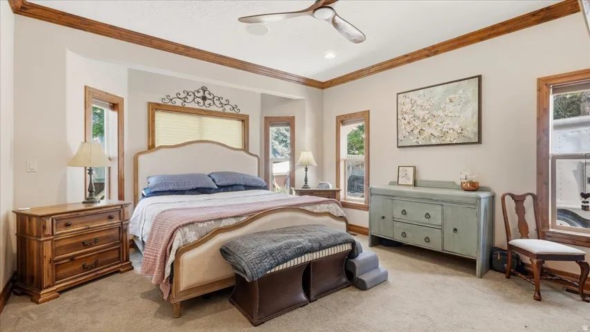 Spacious sleeping area featuring warm wood trim, multiple windows, a ceiling fan, and neutral-toned carpeting