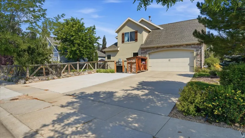 Welcoming driveway and entry featuring a spacious concrete approach, two-car garage with neutral-toned paneling, and mature landscaping with established trees and shrubs framing the property