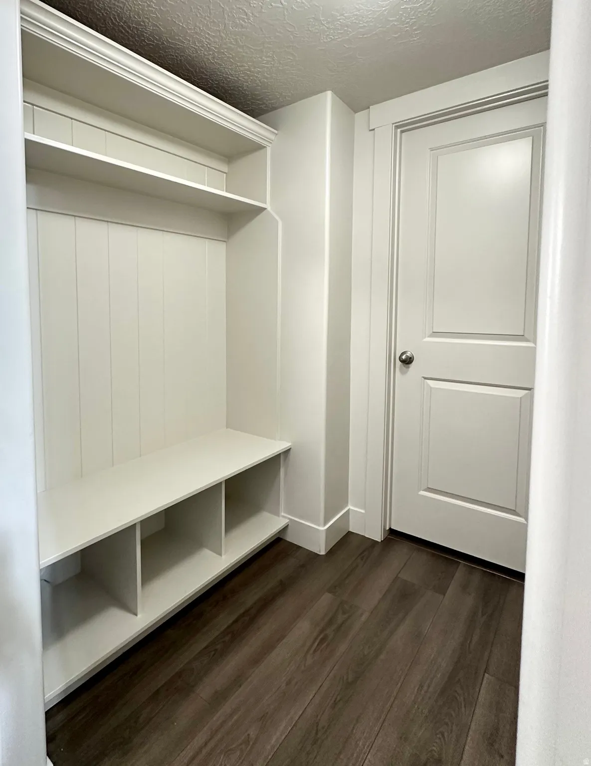Built-in mudroom bench with three storage cubbies, upper shelving, and beadboard paneling, set against wood-finish flooring