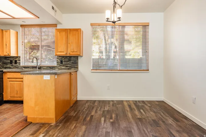 Open-concept kitchen and dining area featuring warm wood cabinetry with ample storage, stainless steel sink, dark tile backsplash, and wood-finish flooring throughout