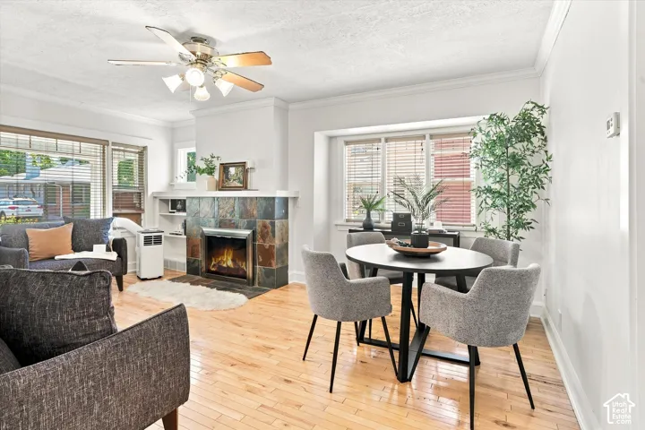 Open-concept living area featuring hardwood flooring, a wood-burning stove with decorative surround, ceiling fan with light fixture, and abundant natural light from multiple windows with blinds