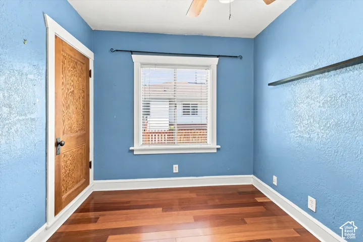 Bright and spacious bedroom showcasing vibrant blue walls, rich wood-finish hardwood flooring, and abundant natural light from a large window with plantation shutters