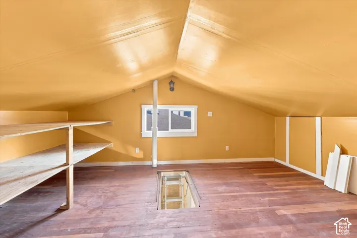Attic space featuring wood-finish flooring, built-in shelving, a window, and exposed ceiling beams