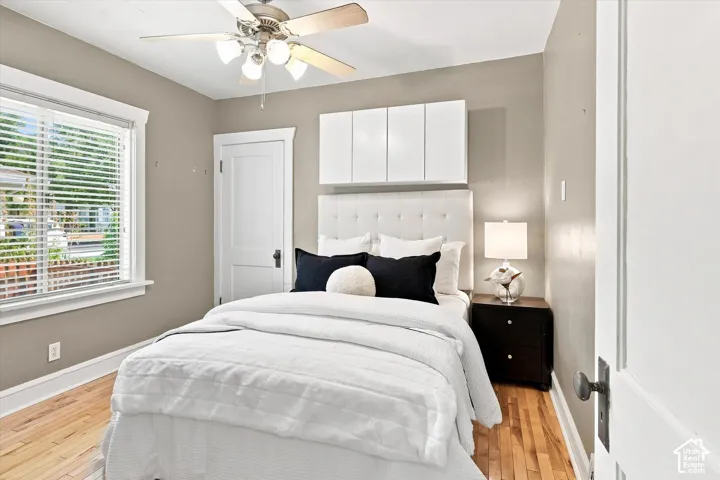Bright bedroom featuring a white upholstered headboard with button tufting, wood-finish flooring, and abundant natural light from a large multi-pane window with plantation shutters