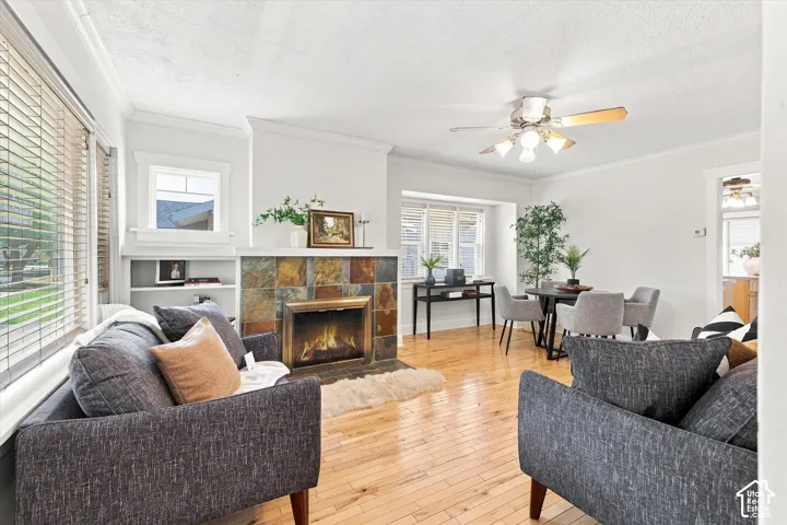 Living area featuring hardwood flooring, a stone-tiled fireplace, built-in shelving, crown molding, and a ceiling fan with light fixture