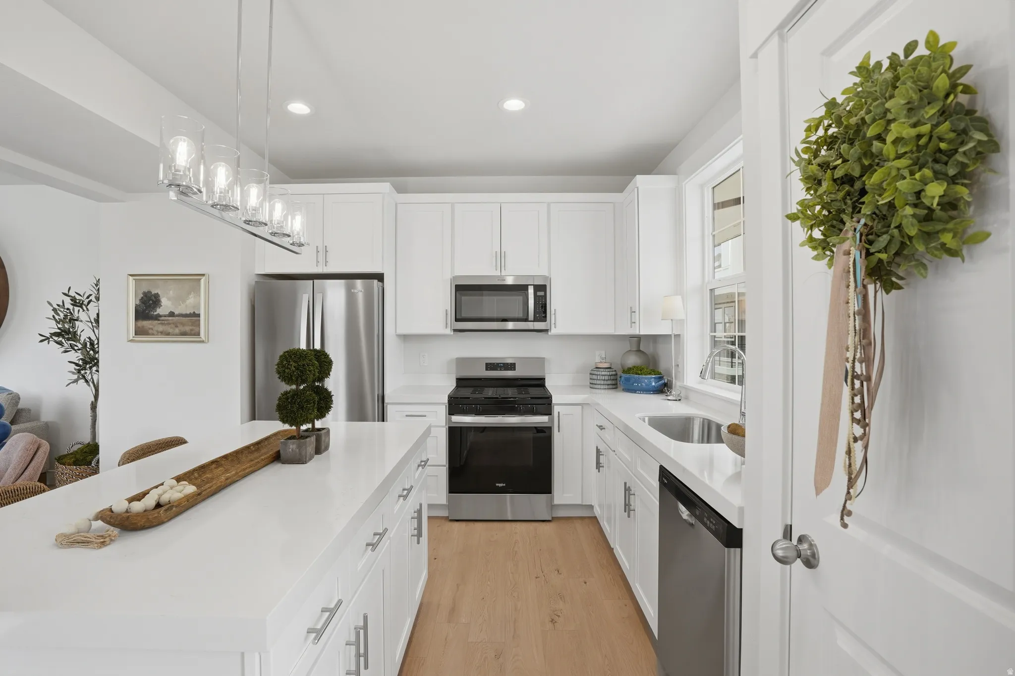 Bright kitchen featuring a substantial island, white cabinetry, integrated stainless steel appliances, wood-finish flooring, and a linear pendant light fixture