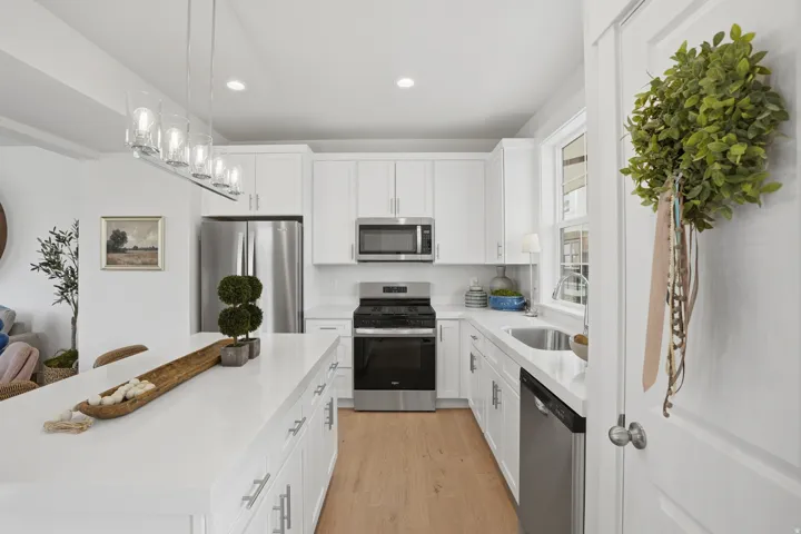 Bright kitchen featuring a substantial island, white cabinetry, integrated stainless steel appliances, wood-finish flooring, and a linear pendant light fixture