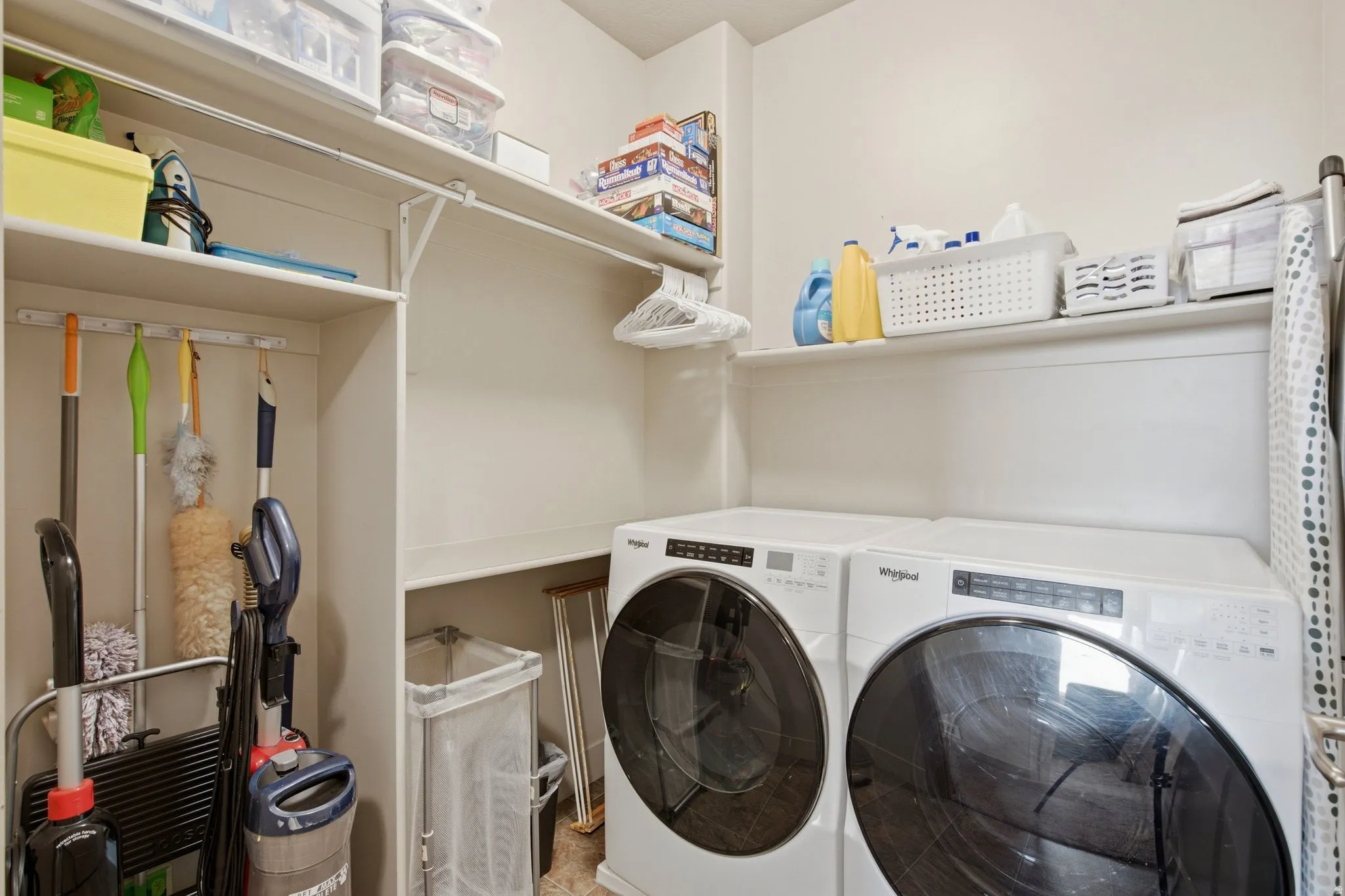 Dedicated laundry room featuring front-load washer and dryer with digital controls, abundant open shelving for storage, wall-mounted utility hooks and rod, and integrated cabinetry for cleaning supplies and household essentials