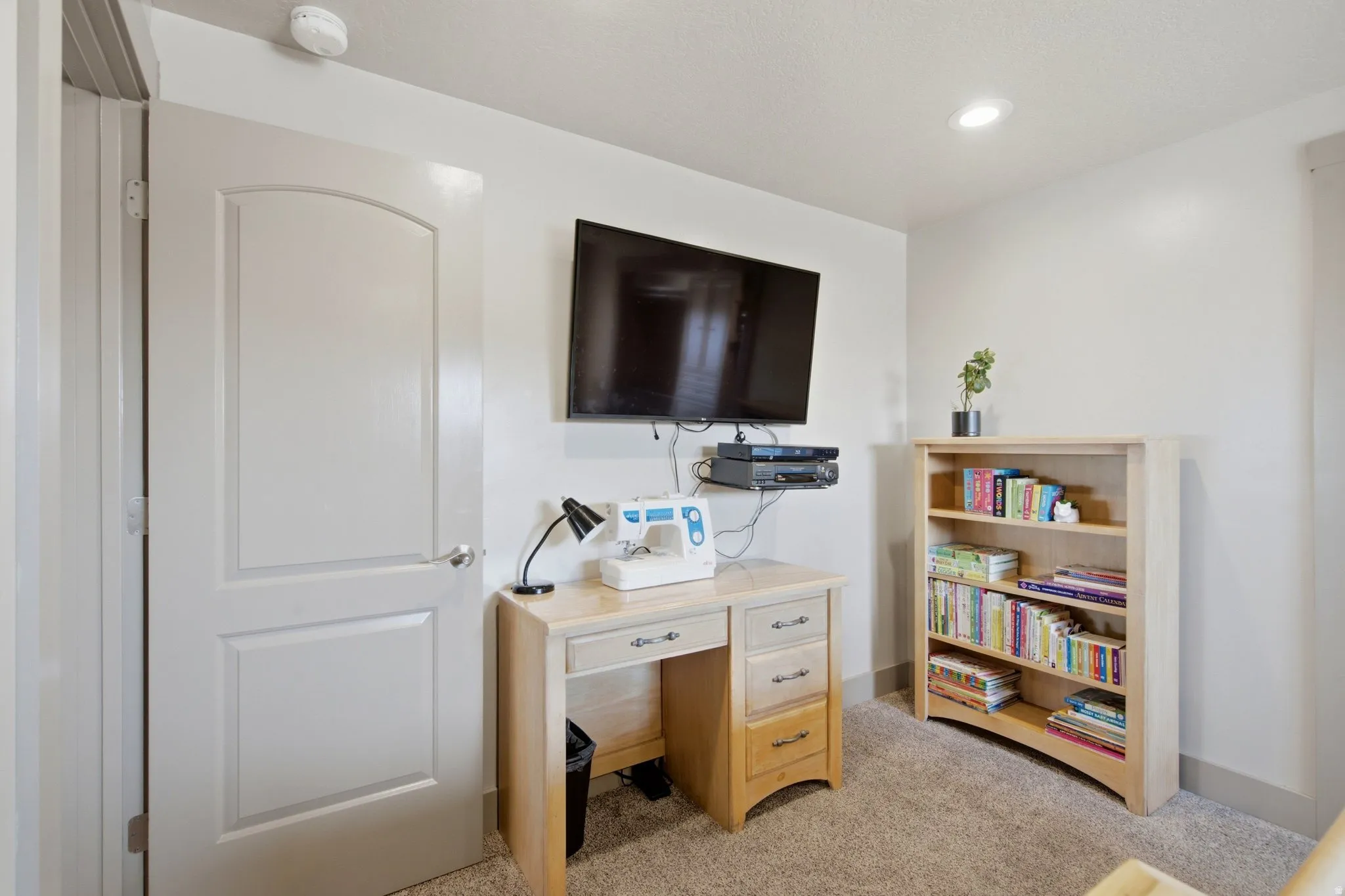 Dedicated home office featuring a wood-finish desk with integrated storage drawers, wall-mounted flat-screen television, recessed ceiling lighting, and a wood-finish open shelving unit displaying books and décor
