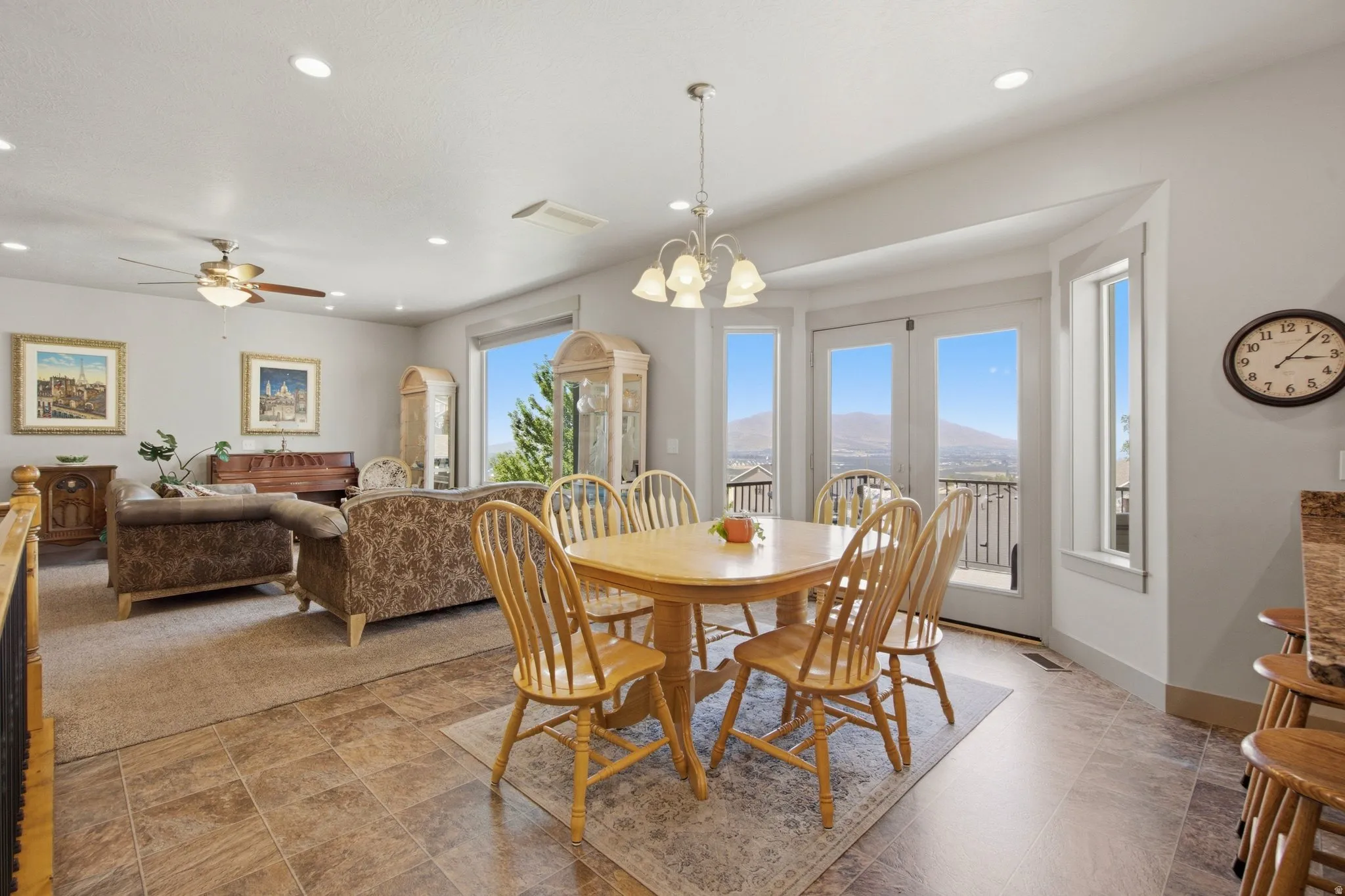 Bright and open living area showcasing expansive glass doors and windows with panoramic vistas, wood-finish flooring throughout, a round dining table with seating for six, upholstered lounge seating, ceiling fan with integrated lighting, recessed ceiling