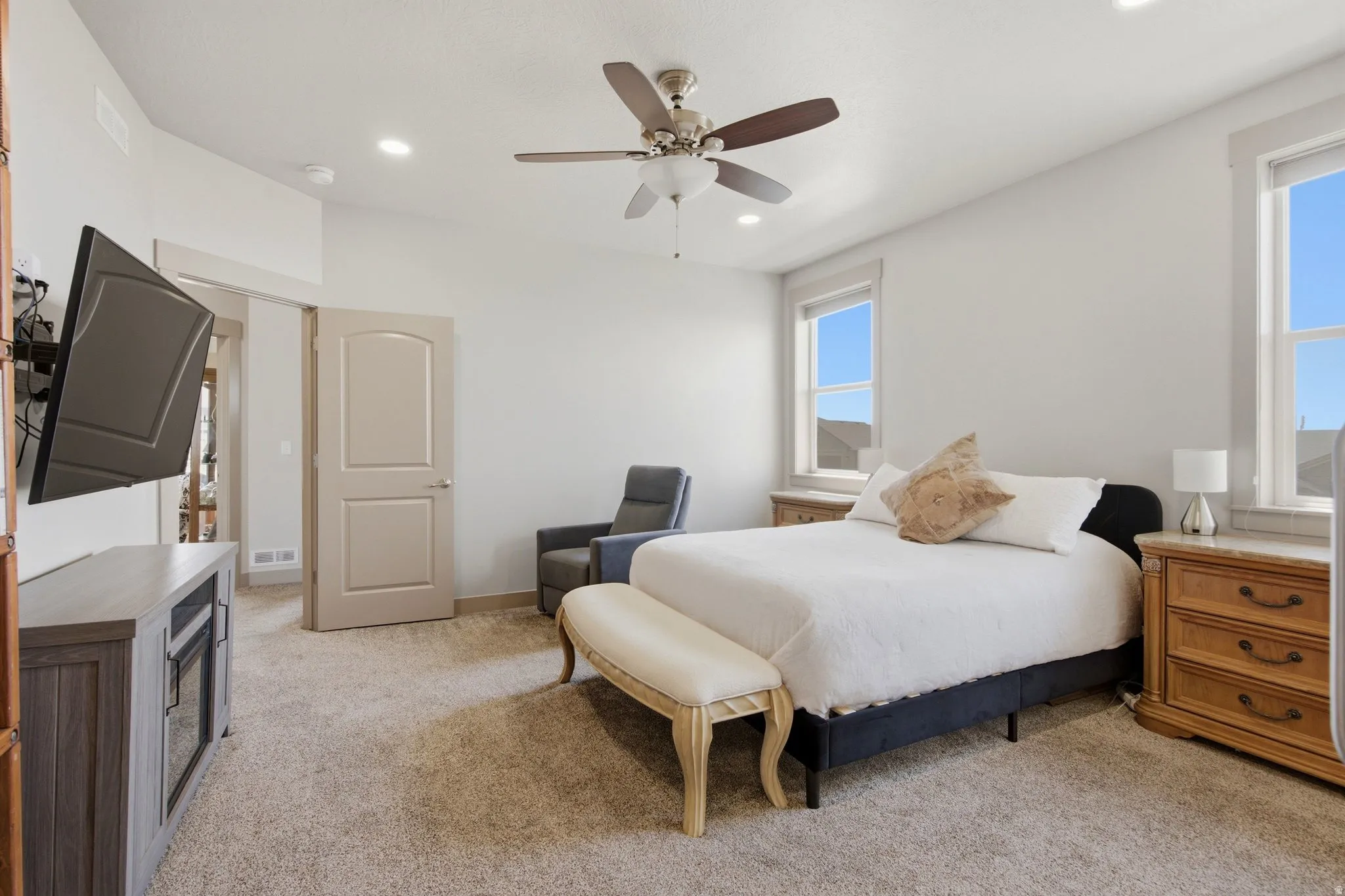 Spacious primary bedroom featuring a wood-frame bed with upholstered base, wood-finish nightstands with drawer storage, ceiling fan with integrated lighting, wall-mounted flat-screen television, and abundant natural light from dual windows