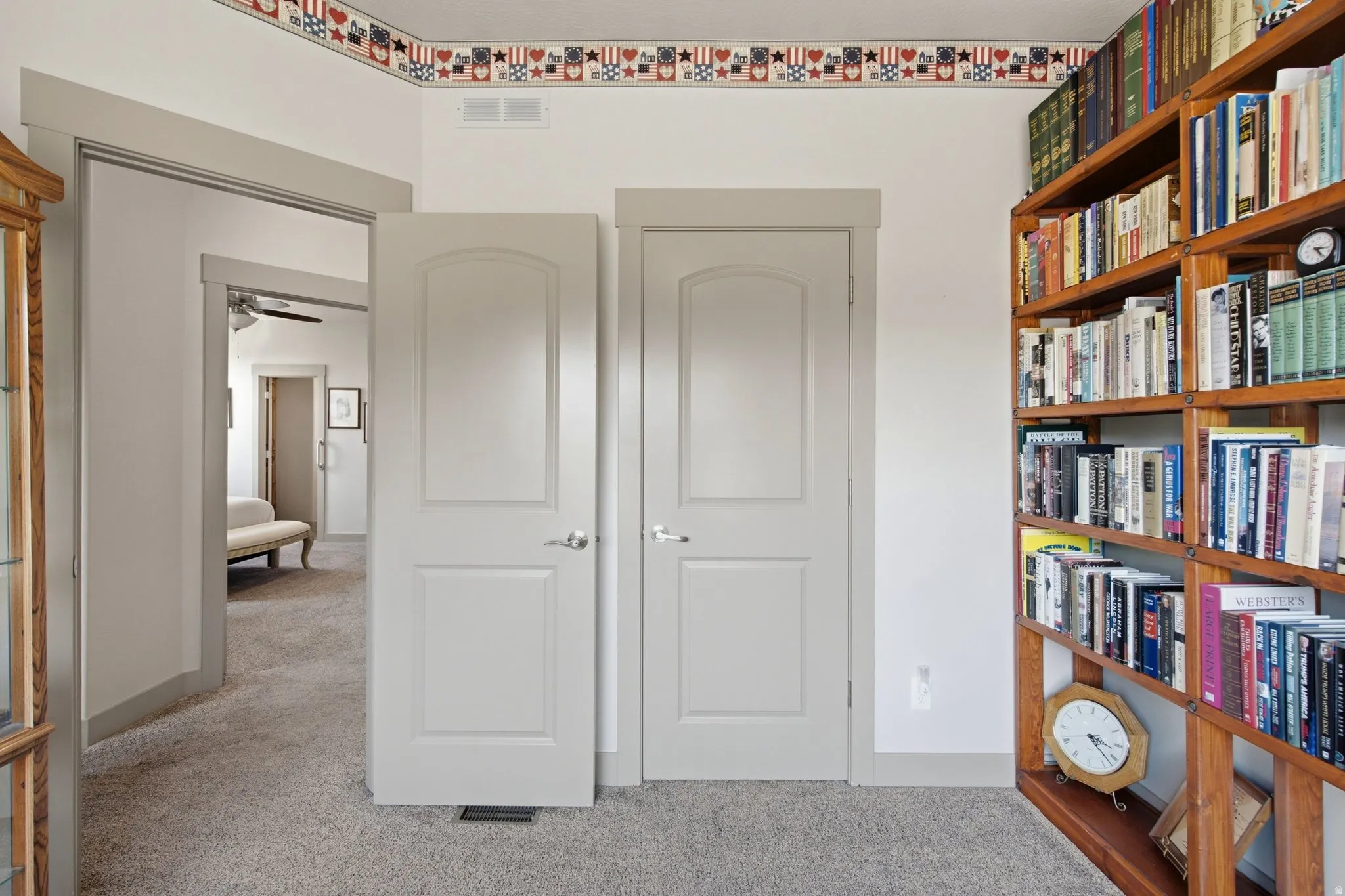 Versatile hallway with elegant white six-panel closet doors, built-in wood-finish shelving displaying curated book collection, neutral carpet flooring, and decorative border detailing along the ceiling