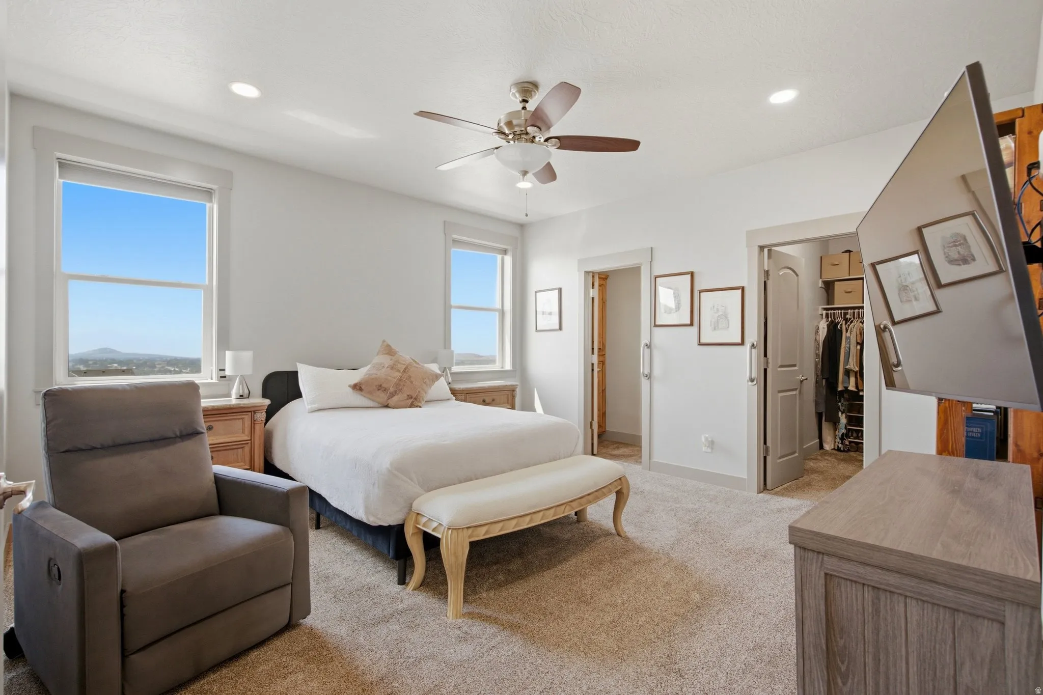 Spacious primary bedroom suite featuring expansive windows with natural light, ceiling fan with integrated lighting, plush carpeting, wood-finish flooring in adjacent areas, built-in closet system, and a wood-framed bench at the foot of the bed