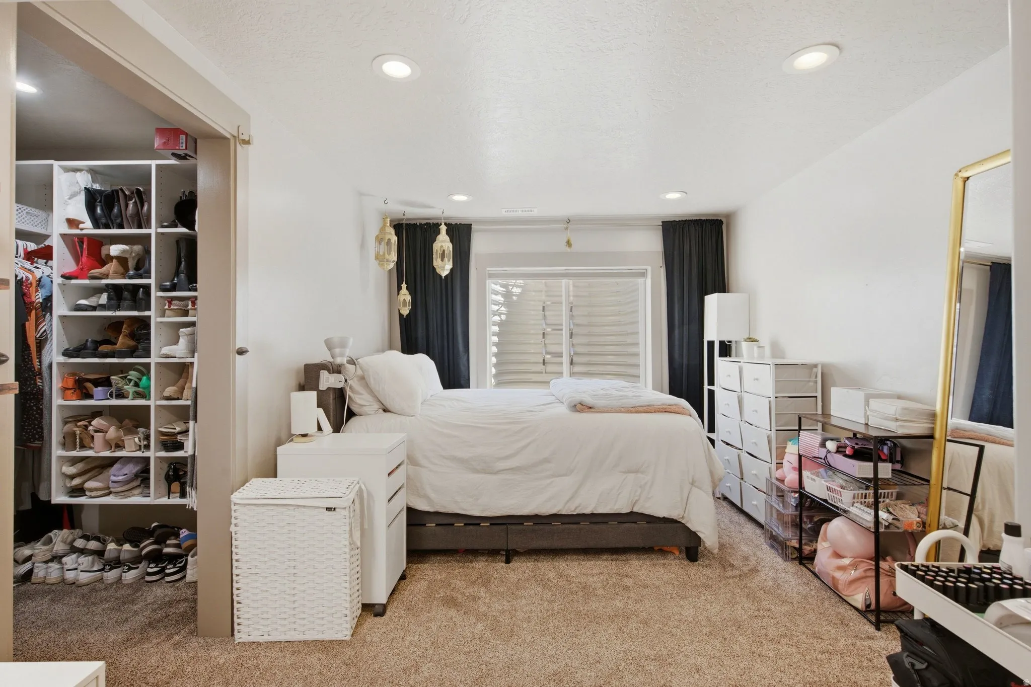Spacious primary bedroom featuring recessed ceiling lighting, dual windows with plantation shutters and blackout drapery, neutral carpeting, and extensive built-in closet storage with shelving and organizational systems