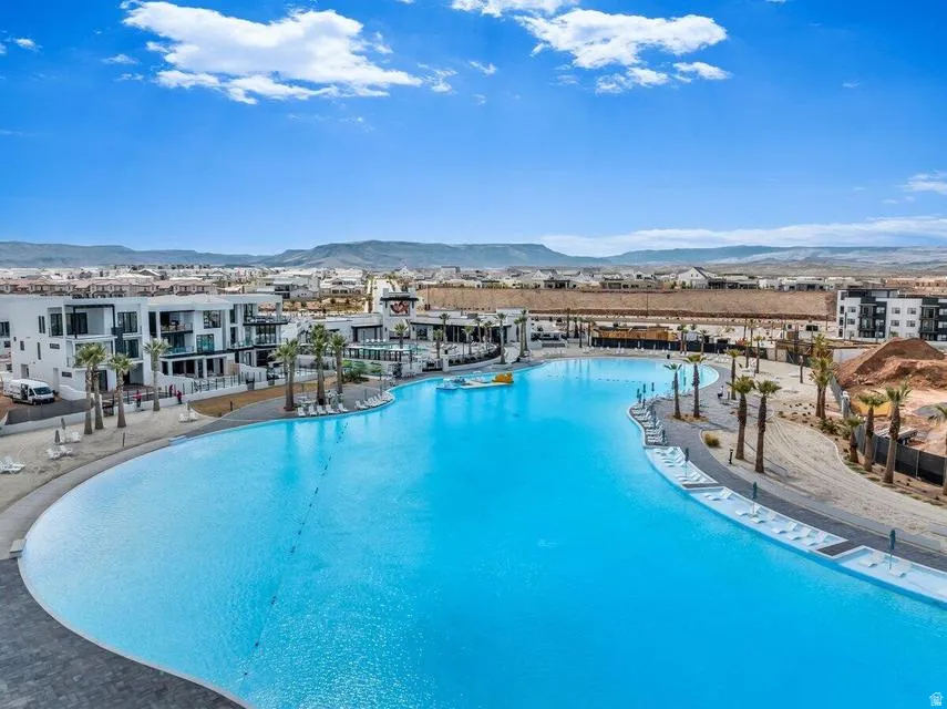 Resort-style lagoon pool featuring expansive crystalline waters, curved sun deck with integrated lounging areas, modern residential buildings with contemporary architecture, and panoramic mountain backdrop