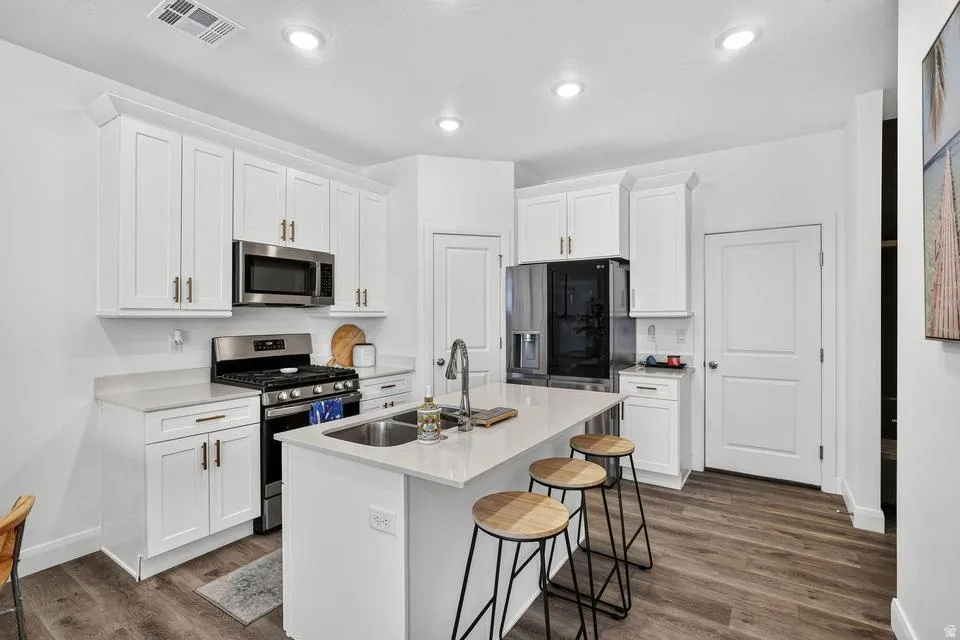 Modern kitchen featuring white shaker-style cabinetry with brushed nickel hardware, stainless steel appliances including a gas range and built-in microwave, quartz countertops, a waterfall-edge island with seating for three, and wood-finish flooring throu