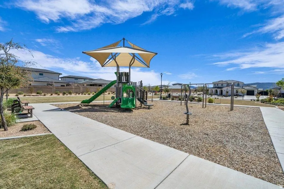 Community playground featuring a multi-level green play structure with slide, shaded canopy, concrete pathways, and gravel surfacing