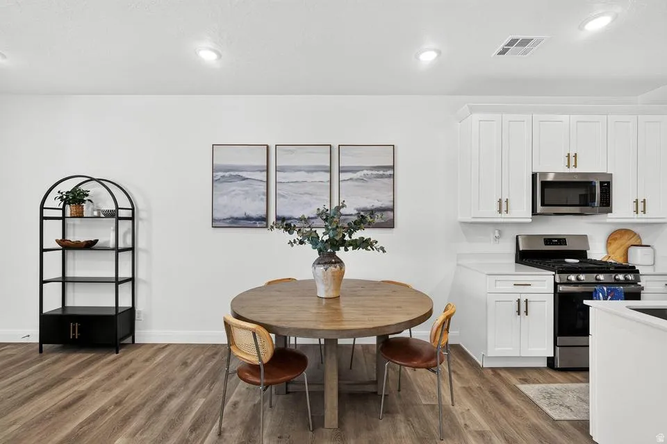 Open-concept kitchen and dining area featuring a round wood-finish dining table, white cabinetry with black hardware, stainless steel appliances including a gas range and microwave, wood-finish flooring throughout, recessed ceiling lighting, and a decorat
