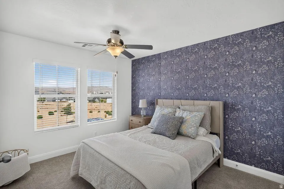 Bright bedroom featuring dual windows with plantation shutters framing expansive natural light, a ceiling fan with integrated lighting, neutral carpeting, and an accent wall with textured wallcovering