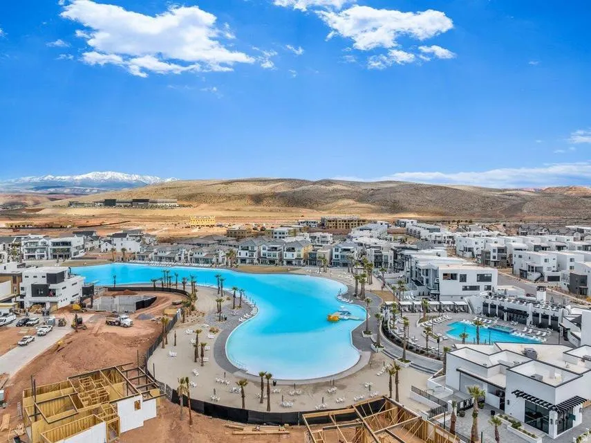 Expansive resort-style community pool complex featuring multiple luminous blue swimming pools with curved architectural design, surrounding white residential buildings with modern construction, and panoramic desert landscape with mountain vistas in the di