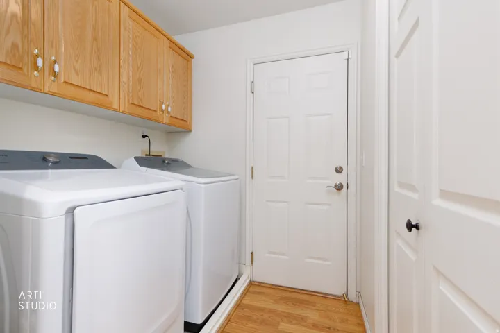 Dedicated laundry room featuring white front-load washer and dryer, wood-finish cabinetry with ample storage, utility sink with bronze faucet, wood-finish flooring, and direct access to the home via white six-panel door