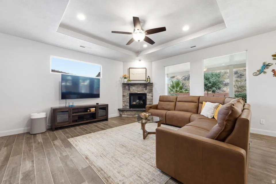 Spacious living area featuring wood-finish flooring, a stone-clad fireplace, and a tray ceiling with recessed lighting and a ceiling fan