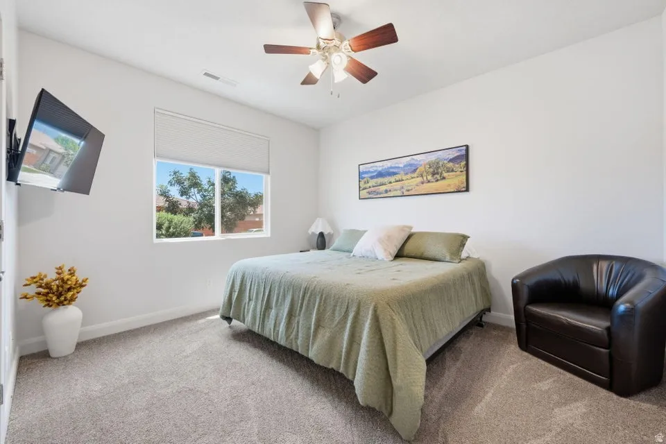 Carpeted interior space featuring a window with a roller shade, a ceiling fan with wood-finish blades, and white walls