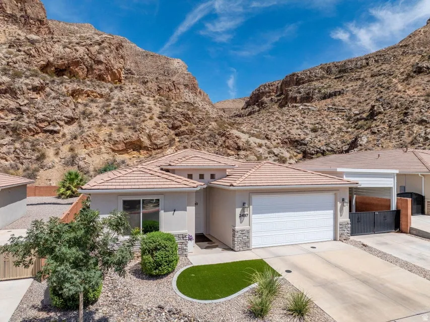 Modern single-story residence nestled against dramatic mountain backdrop, featuring a clean-lined stucco exterior with a pitched tile roof, two-car garage with white overhead door, and a professionally landscaped front yard