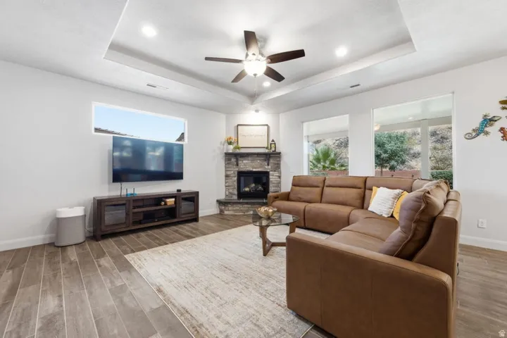 Spacious living area featuring wood-finish flooring, a stone-clad fireplace, and a tray ceiling with recessed lighting and a ceiling fan