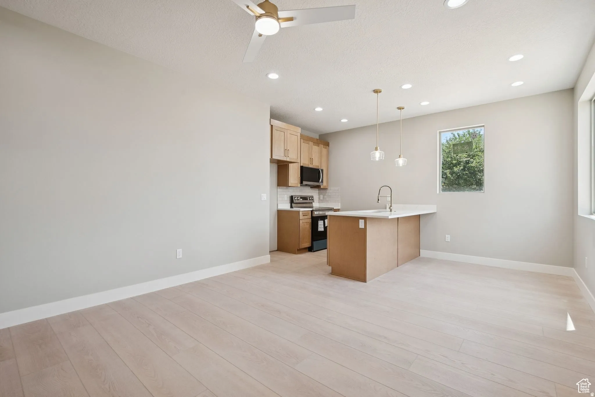 Open-concept kitchen and living area featuring wood-finish flooring throughout, custom wood cabinetry with stainless steel appliances, a centered island with prep space, pendant lighting, recessed ceiling lights, and a ceiling fan