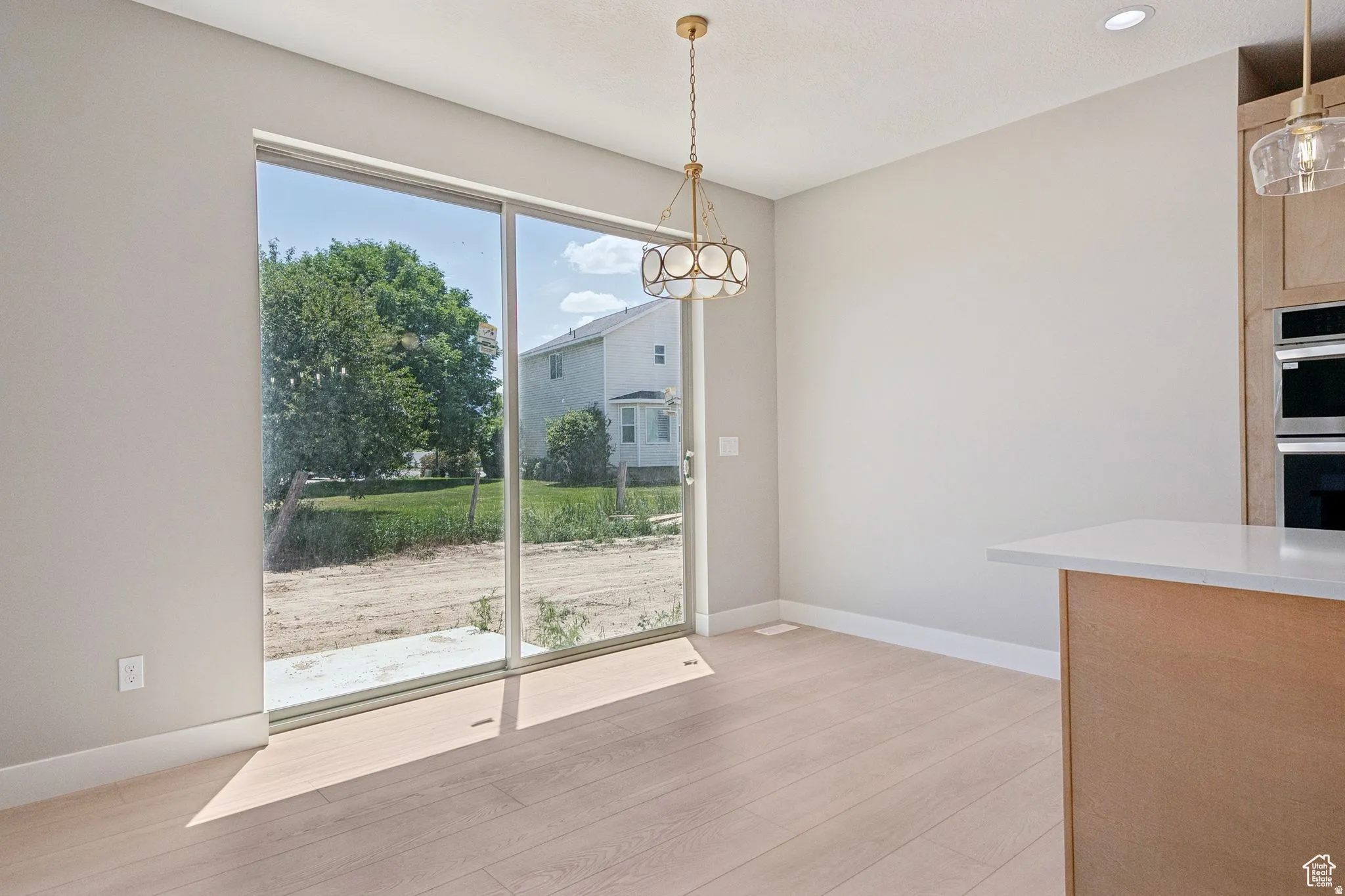 Bright, open dining area featuring expansive sliding glass doors framing natural light, light wood-finish flooring, soft neutral wall finishes, a brass pendant light fixture, and adjacent wood-tone cabinetry with integrated appliances